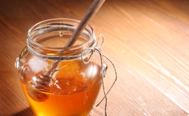 Pot of honey and wooden in it.  Object is on wooden table.